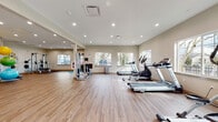 This single-floor fitness center is thoughtfully designed to support a comprehensive range of workout routines in a bright, welcoming environment. The space is finished with durable wood-look flooring throughout, neutral warm-toned walls, and recessed lighting that keeps the area well-illuminated at all times.

The main exercise area is generously proportioned, accommodating multiple zones for different training modalities. A row of large windows lines the perimeter, inviting abundant natural light and providing open views of the surrounding greenery. Cardio equipment is prominently featured, including several treadmills, elliptical trainers, a stationary bike, and a rowing machine, arranged to maximize usable floor space.

A dedicated strength training zone houses a multi-station cable machine, a dumbbell rack with a full range of free weights, and additional floor space for functional training. A well-stocked accessory station offers stability balls, medicine balls, resistance bands, yoga mats, and suspension trainers, catering to stretching and group fitness activities.

A striking floor-to-ceiling glass partition wall separates the fitness area from an adjacent amenity space, creating a sense of openness and visual connectivity. The overall ambiance is clean, modern, and functional, offering an inviting atmosphere well-suited for daily exercise routines.