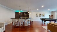 This thoughtfully designed single-floor space offers an impressive blend of entertainment and comfort, unified by rich hardwood flooring and elegant crown molding throughout. The centerpiece of the space is a sleek, fully equipped kitchen and bar area, outfitted with dark wood cabinetry featuring glass-front uppers, a mosaic tile backsplash, granite countertops, built-in stainless steel appliances, and ambient LED accent lighting that adds a modern, vibrant touch. A curved bar counter with upholstered bar seating makes it an ideal gathering spot. Adjacent to the kitchen, a dedicated billiards area provides ample room for a full-size pool table, with framed artwork adorning the walls. The lounge area offers a warm and inviting atmosphere anchored by a stacked stone fireplace with a wall-mounted television above, flanked by wall sconces and large windows that invite natural light and outdoor views. Comfortable seating is arranged around the fireplace, creating a welcoming focal point. A separate dining nook provides an intimate setting for smaller gatherings, complete with a classic wood dining table, tufted upholstered chairs, and decorative artwork. The space also includes a workstation area near sliding glass doors that open to an outdoor patio, enhancing connectivity between the interior and exterior.