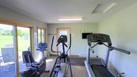 This single-floor fitness room provides a practical and well-equipped space for cardiovascular workouts. The room features a treadmill, an elliptical trainer, and a stationary bike, offering a variety of cardio options. A wall-mounted flat-screen television is positioned for convenient viewing during workouts. The space is finished with neutral green-gray walls, a textured ceiling with fluorescent lighting, and carpeted flooring. Natural light enters through two windows along one wall and a glass-paneled door, which also provides direct exterior access. A baseboard heater runs along the far wall, ensuring comfort during cooler months. The overall layout is functional and straightforward, making efficient use of the available space while keeping equipment accessible. The room's design emphasizes utility, making it a convenient amenity for fitness-focused individuals.