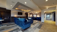 Located in Beavercreek, Ohio, this single-floor community clubroom offers a thoughtfully designed gathering space with a cohesive modern aesthetic. The main lounge area serves as the centerpiece, featuring plush navy blue sectional sofas and accent chairs arranged around a floor-to-ceiling dark tile fireplace topped with a wall-mounted flat-screen television. A coffered ceiling with warm ambient lighting adds architectural depth and a refined atmosphere to the space. Patterned carpet tiles in neutral tones ground the seating area, complementing the room's cool, contemporary color palette. Natural light filters in through large windows, brightening the lounge and offering views of the surrounding community. Adjacent to the main lounge, a dedicated workstation area provides high-top seating along a counter with additional mounted televisions, making it suitable for both casual socializing and focused tasks. The kitchenette area is outfitted with dark cabinetry, stone countertops, a sink, a mosaic tile backsplash, pendant lighting, and vibrant blue bar stools, offering a functional yet stylish refreshment space. Decorative wall art adds personality throughout. Separate men's and women's restrooms are conveniently accessible, accented by a tall potted plant that adds a natural touch to the corridor.
