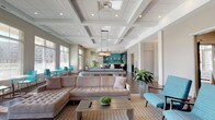 This well-appointed single-floor amenity clubhouse offers a thoughtfully designed space for socializing and recreation. The layout centers around a spacious open-concept lounge and billiards area, anchored by a sleek pool table beneath a striking coffered ceiling with recessed lighting. Rich dark hardwood flooring runs throughout, complementing the neutral, contemporary color palette accented by teal and aqua furnishings.

Floor-to-ceiling windows line the perimeter, inviting abundant natural light and offering direct views of an outdoor pool and surrounding community. Glass doors provide seamless access to the pool deck, enhancing the indoor-outdoor connection. The lounge area features a generous tufted sectional sofa, a patterned area rug, and casual seating arrangements ideal for gathering.

The catering kitchen is a standout feature, equipped with dark wood cabinetry, white countertops, dual under-counter beverage refrigerators, and a vibrant teal subway tile backsplash that adds a bold design statement. A pass-through window connects the kitchen to the main lounge area.

A separate media nook offers a more intimate setting, complete with a linear stone-clad fireplace, wall-mounted flat-screen television, and tufted accent chairs. The overall ambiance is modern, bright, and inviting, making this clubhouse a versatile and stylish community gathering space.