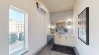 This rooftop terrace and supporting kitchenette area offers a compelling combination of indoor utility and expansive outdoor living. The interior kitchenette is cleanly designed with light wood cabinetry, a built-in beverage refrigerator, a countertop sink, and open floating shelves backed by a soft grey subway tile accent wall. A wall-mounted mini-split unit ensures climate comfort, and a window provides natural light with views to the surrounding cityscape.

The rooftop terrace itself is the standout feature of this space. Finished with richly toned teak deck tiles arranged in a checkerboard pattern, the outdoor area is thoughtfully organized into multiple seating and dining zones. A lounge area features rope-wrapped chairs and a sofa paired with a stone drum coffee table, ideal for relaxed gatherings. Nearby, multiple dining tables with coordinating chairs accommodate larger groups in a more formal setting.

The perimeter is defined by a clean horizontal wood privacy wall, framing sweeping panoramic views of the surrounding urban landscape, lush tree canopies, and distant mountain ridgelines under open sky. The terrace receives abundant natural light throughout the day, making it a versatile and inviting amenity space suited for both social events and quiet retreat.