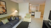 This single-floor space showcases an open-concept layout that seamlessly connects the living area to the kitchen, creating a cohesive and functional environment. The living area is carpeted and features a charcoal sofa accented with patterned throw pillows, a cream armchair with a green cushion, and a sleek dark coffee table, all arranged around a central seating area. A side table with a white lamp adds warm ambient lighting to the space. The accent wall in a muted sage green provides a subtle contrast to the surrounding cream-toned walls, lending depth and visual interest to the room. A ceiling fan is mounted overhead, contributing to the space's practicality and comfort. The kitchen, visible from the living area, features white cabinetry, light countertops, and standard appliances including a dishwasher, all finished in a clean, neutral palette. The open sightline between the two areas enhances the sense of spaciousness. Overall, the space presents a calm, well-organized atmosphere with a cohesive neutral color scheme suited for everyday living.