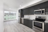 This single-floor unit showcases a clean, contemporary design throughout, with light hardwood-style flooring and crisp white walls creating a bright, cohesive aesthetic. The open-plan layout connects the kitchen and living area seamlessly, maximizing the sense of space and encouraging easy movement between zones.

The kitchen is well-appointed with sleek grey high-gloss cabinetry, stainless steel appliances including a refrigerator, dishwasher, electric range, and microwave, along with a white subway tile backsplash and light quartz countertops. A stainless undermount sink with a modern pull-down faucet adds a polished finishing touch.

The living area benefits from a large picture window that draws in abundant natural light and offers views of the surrounding neighborhood. Recessed lighting supplements the natural light throughout.

The unit includes two bathrooms. The full bathroom features marble-style large-format wall tiles surrounding a soaking tub, paired with wood-look flooring. The second bathroom offers a vanity with dark cabinetry, a light countertop, and matching flooring, providing a functional and stylish secondary space. Throughout, the unit maintains a consistent modern and well-finished character.