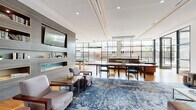 This single-floor space exemplifies contemporary design through its open, flowing layout and carefully curated aesthetic. Floor-to-ceiling steel-framed windows dominate the exterior walls, flooding the interior with natural light and providing views of an adjoining outdoor terrace. Warm hardwood flooring extends throughout, grounding the space and providing visual continuity across its distinct zones.

The main gathering area features a generously sized lounge with plush, neutral-toned seating arranged atop a richly patterned blue area rug. A textured stone-clad accent wall houses built-in shelving and a wall-mounted flat-screen television, adding architectural depth. Elegant bronze-toned vertical slatted partitions serve as stylish room dividers, creating a sense of separation between zones while maintaining an open, airy feel.

A central kitchen island with a dark stone countertop anchors the culinary area, complemented by warm wood cabinetry and pendant lighting suspended from above. Adjacent to the kitchen, a formal dining configuration offers ample seating beneath a linear chandelier.

A dedicated game room with a full-size billiard table, additional display screens, and bar-height seating rounds out the amenities, making this a highly versatile and refined communal space.