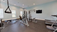 Located at 82 Ardmore Gardens Dr in Hardeeville, South Carolina, this fitness center offers a well-equipped, single-floor space designed to support a full range of workout routines. The interior is finished with soft blue-gray walls and crisp white trim, creating a clean and inviting atmosphere. Durable rubber flooring throughout provides both safety and comfort underfoot.

The facility is comprehensively outfitted with a variety of strength-training machines, including multi-function cable systems, selectorized weight machines, and a functional trainer with adjustable cable attachments. A dedicated free weights area features a dumbbell rack with a full range of weights, along with flat and adjustable benches. For cardio, the space includes multiple treadmills, elliptical trainers, and stationary bikes, several equipped with individual display screens.

A designated functional training zone offers interlocking floor mats, medicine balls with a storage rack, and a stability ball, ideal for flexibility and core work. A wall-mounted flat-screen television adds entertainment for users during workouts. Recessed lighting and multiple windows with slatted blinds allow natural light to fill the space while maintaining privacy. The overall layout is thoughtfully organized to maximize usable floor space and ensure smooth traffic flow throughout the facility.