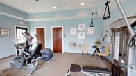 This fitness center offers a thoughtfully designed workout environment spread across a single floor. The space is finished with soft blue walls and white tray ceilings, creating a calm and motivating atmosphere. Recessed lighting and ceiling fans ensure the space remains well-lit and comfortable during use.

The cardio area is equipped with multiple treadmills and an elliptical trainer, positioned near large windows that allow ample natural light and views of the surrounding greenery. A wall-mounted flat-screen television provides entertainment during workouts. On the strength training side, the space features a multi-functional cable machine, a rowing machine, a weight bench, and a rack of free weights including dumbbells and barbells, catering to a full range of exercise routines.

A dedicated hydration station with a dual drinking fountain is conveniently mounted on the wall, alongside seating for rest between sets. Decorative coastal-themed artwork adds a welcoming touch to the interior. The fitness center also includes restroom access through solid wood doors, a sanitation station, and waste receptacles, reflecting attention to cleanliness and user convenience. Natural light flows in through multiple windows, enhancing the open and airy feel of the space throughout the day.