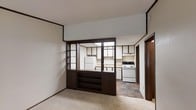 This single-floor unit presents a compact yet functional layout, as revealed by the overhead floor plan and interior imagery. The space is organized into distinct living and kitchen zones, connected by an open, flowing arrangement that maximizes the available square footage.

A standout architectural detail is the bold, dark wood-framed partition that separates the main living area from the kitchen. This divider features open shelving on the lower half and glass-panel sections above, allowing sightlines between the two spaces while still providing a sense of definition. The shelving unit offers practical built-in storage and display space.

The living area is finished with light-toned carpet, contributing to a neutral and understated aesthetic. In contrast, the kitchen features a slate-toned tile floor, providing a durable, easy-to-clean surface. The kitchen is equipped with white appliances, including a freestanding electric range and a full-sized refrigerator, alongside upper and lower cabinetry trimmed with dark wood accents that echo the divider's design.

Natural light enters through windows visible in the floor plan, and the white walls throughout help keep the interior feeling bright. The overall ambiance is modest and functional, with a retro character defined by the dark wood detailing.