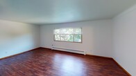 This single-floor unit offers a practical and straightforward layout across several rooms, all unified by rich, dark hardwood-style flooring and crisp white walls. The living area is generously proportioned, featuring a wide window that frames views of surrounding greenery and allows natural light to fill the space. A wall-mounted air conditioning unit provides additional climate control, complementing the baseboard heating system found throughout the unit.

Two bedrooms mirror a similar aesthetic — each room features a ceiling fan with integrated lighting, a window overlooking lush tree canopy, and baseboard heating positioned beneath the sill. The clean white walls and consistent flooring create a cohesive feel between spaces.

The kitchen is efficiently arranged, equipped with a white electric coil range, a full-size refrigerator, and warm oak-toned upper cabinetry providing ample storage. A range hood is mounted above the stove, and the layout allows easy movement between the cooking and adjacent areas.

The bathroom features tile wainscoting along the walls, a white vanity with a single basin sink, a mirrored medicine cabinet with wood framing, and a tub and shower combination. A globe light fixture above the mirror provides functional illumination. The overall ambiance of the unit is clean, neutral, and move-in ready.