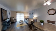This single-floor residence offers a thoughtfully arranged open-concept layout that seamlessly connects the living and dining areas into one cohesive, functional space. The living area is centered around a light gray sofa paired with a patterned accent chair, anchored by a woven area rug and a rustic wood-and-metal coffee table. Soft ambient lighting from table lamps creates a warm, inviting atmosphere throughout. A large sliding glass door allows natural light to fill the room while providing a view to the exterior, dressed with sheer curtains that diffuse the light gently across the space. The walls are painted in a calming light blue-gray tone, complementing the crisp white ceiling and contributing to a clean, modern aesthetic. Rich dark hardwood-style flooring runs throughout, adding warmth and contrast to the lighter color palette. The dining area, positioned adjacent to the living space, features a round wooden dining table with matching chairs, offering an intimate setting for meals. Framed artwork displayed on the accent wall adds a decorative touch that ties the overall design together. The space presents a balanced combination of comfort, style, and practicality suitable for everyday living.