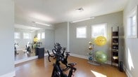 Located at 4531 Langdon Rd in Morrisville, NC, this single-floor fitness studio is thoughtfully designed to support a well-rounded exercise routine. The space features a clean, modern aesthetic with light gray walls, white trim, and warm wood-tone flooring that creates an inviting and energizing atmosphere. Multiple spin bikes are arranged to allow ample room for movement, making the area well-suited for cycling workouts. A large floor-to-ceiling mirror lines one wall, enhancing the sense of space while allowing users to monitor their form during exercise. A freestanding rack holds a variety of exercise balls in multiple sizes and colors, providing versatile options for strength and flexibility training. Natural light flows into the room through several white-framed windows, offering views of the surrounding greenery and keeping the environment bright and welcoming. Overhead fluorescent lighting ensures consistent illumination throughout the space. The layout is open and uncluttered, prioritizing functionality and ease of movement. This fitness studio is well-appointed for both cardio and functional training, making it a practical and comfortable space for daily workouts.