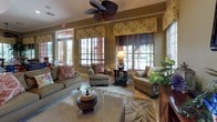 This single-story space showcases a consistently styled interior with a warm, tropical aesthetic carried throughout each area. The living room serves as a central gathering space, featuring tile flooring topped with a botanical-patterned area rug, crown molding, and a decorative ceiling fan. Multiple windows dressed with wooden blinds and coordinating floral valances allow natural light to filter in while offering views of lush greenery outside. Comfortable upholstered seating is arranged around a coffee table, creating an inviting atmosphere.

An adjoining dining nook continues the tropical theme, with a round dark-wood table surrounded by cane-back chairs with cushioned seats. A large potted tropical plant anchors a corner surrounded by windows, enhancing the garden-like ambiance. A matching ceiling fan completes the room.

The kitchen is well-equipped with rich dark-wood cabinetry, a dark granite countertop island with an undermount sink, and stainless steel appliances. Pendant lighting illuminates the workspace, while woven rattan barstools add a natural texture at the island. Decorative vessels and greenery displayed atop the upper cabinets tie the space together with the home's overall tropical character.