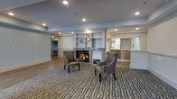 This single-floor space offers a warm and inviting atmosphere centered around a well-appointed common lounge area. The focal point of the room is a classic fireplace framed by a built-in shelving unit with open display niches, providing both functional storage and a refined aesthetic. Two high-back upholstered accent chairs are positioned facing the fireplace, creating an intimate seating arrangement ideal for relaxation or conversation. The flooring transitions between smooth hardwood-style planks and a richly patterned area rug that anchors the seating area, adding texture and visual interest to the space. Recessed lighting is distributed evenly across the ceiling, complementing the clean, neutral color palette of whites and soft grays that defines the overall design. Crown molding details along the upper walls contribute a polished, finished look throughout. Adjacent to the lounge, a reception or service counter is visible, featuring a textured stone or tile accent wall that introduces a warm, earthy contrast to the otherwise cool tones of the space. A secondary room or corridor is accessible in the background, suggesting a well-connected, multi-use layout. Overall, the space balances comfort and functionality with a clean, contemporary design aesthetic.