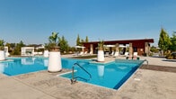 Located at 6650 Liberty Grand Blvd in Powell, Ohio, this resort-style outdoor amenity complex is thoughtfully designed to offer a comprehensive recreational experience. At the heart of the space are two large swimming pools with crystal-clear blue water, surrounded by generous concrete pool decks lined with lounge chairs and patio umbrellas, ideal for sunbathing and relaxation. Large decorative planters with tropical foliage add a resort-like aesthetic to the pool surroundings.

A standout feature is the spacious covered pavilion, constructed with warm wood ceilings and stone columns, housing an outdoor kitchen with grills, a bar area, mounted televisions, ceiling fans, and comfortable seating — all oriented toward the pool for seamless indoor-outdoor living. Adjacent to the pavilion, a well-manicured putting green and a bocce ball court provide recreational options set against vibrant landscaped borders.

Throughout the grounds, lush turf areas interspersed with decorative concrete pathways create distinct lounging zones furnished with contemporary outdoor sofas and chaise lounges. Mature trees and colorful perennial garden beds with ornamental grasses and flowering shrubs frame the perimeter, offering both privacy and natural beauty under the open Ohio sky.