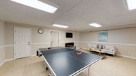 This single-floor amenity space is thoughtfully organized into three distinct functional areas, each designed to serve a specific purpose for its users. The largest area is a recreation room featuring light hardwood-style flooring, a drop-panel ceiling with flush-panel lighting, and neutral-toned walls accented by a painted brick wainscoting detail. A full-size ping-pong table serves as the centerpiece, while a cozy lounge corner offers a sofa, coffee table, wall-mounted flat-screen TV, and a sleek electric fireplace, creating a warm and social atmosphere. An adjacent workstation room provides a quieter retreat, furnished with a built-in desk, computer setup, and ergonomic seating, finished with patterned carpet tile flooring and soft grey walls. The fitness center occupies a generously sized room fitted with the same carpet tile flooring and drop-panel ceiling. It is well-equipped with multiple treadmills, elliptical trainers, a stationary recumbent bike, and an upright cycle, all arranged for easy access. Wall-mounted monitors are positioned throughout for entertainment during workouts. Together, these three zones offer a comprehensive and well-maintained amenity suite suited for recreation, productivity, and physical fitness.