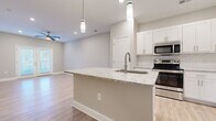 Located at 8000 Carlow Dr in Kernersville, NC, this single-floor, one-bedroom, one-bathroom residence offers a thoughtfully designed open-concept layout with modern finishes throughout. The combined kitchen and living area serves as the heart of the home, centered around a granite-topped kitchen island with an integrated sink and stylish pendant lighting above. White shaker-style cabinetry, a classic subway tile backsplash, and a full suite of stainless steel appliances — including a refrigerator, range, and microwave — complete the kitchen. Light hardwood-style flooring extends across the main living areas, complemented by neutral wall tones and clean white baseboards that lend a fresh, cohesive feel. The living area benefits from natural light through French doors and a ceiling fan for year-round comfort. The spacious bedroom features soft carpeting, a ceiling fan, and a window with blinds that invites natural light. A dedicated closet provides practical storage. The full bathroom is finished with a granite-top vanity, white cabinetry with brushed gold hardware, and tile flooring, creating a clean and polished look. Hallways connect the spaces efficiently, completing this well-appointed single-level home.
