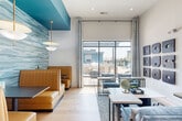 Located at 112 Point Pl in Mooresville, NC, this thoughtfully designed single-floor space serves as a vibrant community lounge and workspace. The interior blends coastal-inspired aesthetics with modern sophistication through a cohesive palette of teal, warm gold, and crisp white tones throughout.

The living area is the heart of the space, boasting soaring ceilings that amplify its openness. Large floor-to-ceiling windows flood the room with natural light and offer views of an outdoor pool area. Rich light oak hardwood flooring runs throughout, complementing plush blue velvet seating arranged around a marble-topped coffee table. A striking textured wave-panel accent wall in teal frames a dining nook outfitted with tan leather booth seating, dark bistro tables, and elegant gold pendant lighting.

The two office spaces offer a more focused atmosphere. Each features a sleek marble-surface desk, a cognac leather chair, and refined geometric patterned wallcovering, creating a calm and productive environment. Soft overhead pendant lighting and lush greenery add warmth to each workstation.

Curated artwork, including a bold grid of abstract panels, adds a gallery-like character to the lounge. The seamless blend of functional zones and design-forward details makes this space both practical and visually compelling.