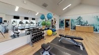This fitness center offers a comprehensive workout environment designed to accommodate a wide range of exercise routines. The space is finished with light hardwood-style flooring throughout, contributing to a clean and energetic atmosphere. Vaulted ceilings in the strength training area add a sense of openness, while flush linear lighting ensures consistent, well-distributed illumination across the room.

The cardio zone is lined with large windows that invite natural light and provide views of surrounding trees, creating a refreshing backdrop during workouts. This area is equipped with multiple treadmills, elliptical machines, and a stationary bike, each accompanied by wall-mounted flat-screen televisions for entertainment.

The free weights and functional training section features a full dumbbell rack, adjustable benches, stability balls in varying sizes, medicine balls, resistance bands, and foam rollers neatly organized on multi-tier storage racks. A large wall mirror enhances the sense of space and supports proper form during exercise.

A standout design element is the floor-to-ceiling nature-inspired mural depicting a misty forest scene, adding a distinctive and calming visual character to the space. A ceiling fan complements the ventilation system, completing this thoughtfully appointed fitness facility.
