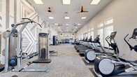 Located at 701 Watkins View Drive in Midlothian, Virginia, this fitness center offers a generously sized, single-floor exercise space designed to support a comprehensive workout experience. The room is characterized by soaring ceilings that enhance the open, airy atmosphere, complemented by an array of large windows along one wall that flood the interior with natural light and provide views of the outdoor surroundings, including a glimpse of a pool area beyond.

The layout is thoughtfully organized into distinct zones. A dedicated cardio section lines the windowed wall and features a row of treadmills, elliptical trainers, and air bikes, including Assault Fitness and other commercial-grade machines. Adjacent to this area, a strength and free weights zone includes multiple racks of dumbbells, barbells, and a cable machine, offering options for a full range of resistance training.

A large open floor area provides space for stretching, functional training, or group exercise. Rubber flooring and neutral carpet tiles delineate the activity zones, while multiple ceiling fans ensure consistent airflow throughout. A striking geometric-patterned accent wall adds a bold visual element to the otherwise clean, modern aesthetic. Built-in shelving, wall-mounted TVs, and coat hooks along one wall contribute to the room's practicality and polished finish.