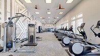 Located at 701 Watkins View Drive in Midlothian, Virginia, this fitness center offers a generously sized, single-floor exercise space designed to support a comprehensive workout experience. The room is characterized by soaring ceilings that enhance the open, airy atmosphere, complemented by an array of large windows along one wall that flood the interior with natural light and provide views of the outdoor surroundings, including a glimpse of a pool area beyond.

The layout is thoughtfully organized into distinct zones. A dedicated cardio section lines the windowed wall and features a row of treadmills, elliptical trainers, and air bikes, including Assault Fitness and other commercial-grade machines. Adjacent to this area, a strength and free weights zone includes multiple racks of dumbbells, barbells, and a cable machine, offering options for a full range of resistance training.

A large open floor area provides space for stretching, functional training, or group exercise. Rubber flooring and neutral carpet tiles delineate the activity zones, while multiple ceiling fans ensure consistent airflow throughout. A striking geometric-patterned accent wall adds a bold visual element to the otherwise clean, modern aesthetic. Built-in shelving, wall-mounted TVs, and coat hooks along one wall contribute to the room's practicality and polished finish.