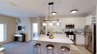 Located at 1910 Capital Creek Dr in Wake Forest, NC, this single-floor residence offers a well-designed open-concept layout that seamlessly connects the kitchen and living areas. The kitchen is a standout feature, showcasing white shaker-style cabinetry, a classic subway tile backsplash, and light-toned marble-look countertops that lend a clean, modern aesthetic. A spacious central island with seating for multiple guests serves as both a functional workspace and a social hub, illuminated by a striking linear pendant light fixture with glass shades that adds a touch of contemporary elegance. Stainless steel appliances, including a range and microwave, complete the kitchen's polished, professional look. The open flow into the living area is enhanced by neutral-toned carpeting and soft, neutral wall colors that create a calm and inviting ambiance. Built-in shelving provides decorative display space, adding character to the common areas. The layout allows natural light to move freely through the space, contributing to a bright and airy atmosphere throughout. A glimpse into the adjacent bedroom reveals continued use of clean, neutral finishes, maintaining a cohesive design language across the entire residence.