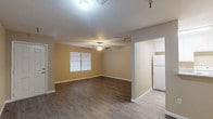 This single-story unit offers a thoughtfully arranged layout with a consistent, neutral color palette of warm beige tones and crisp white trim throughout. The living area is generously proportioned, featuring durable wood-look laminate flooring, a ceiling fan with integrated lighting, and a window fitted with blinds that allows natural light to filter in, creating a bright and comfortable atmosphere.

The layout flows into a secondary room of comparable size, also equipped with a ceiling fan and the same wood-look flooring. This space provides access to additional rooms via white paneled doors. A third room, featuring its own ceiling fan, connects to what appears to be a walk-in closet outfitted with a full-width hanging rod and matching flooring — providing practical storage capacity.

The galley-style kitchen is efficiently designed with white cabinetry, granite-patterned countertops, a white electric range, dishwasher, and refrigerator. The layout maximizes counter space along both sides of the kitchen corridor.

The bathroom includes a walk-in tub/shower combination finished in white tile, equipped with multiple stainless steel grab bars for accessibility, along with an overhead cabinet for storage. White baseboards run consistently throughout the unit, contributing to a clean, well-maintained aesthetic overall.