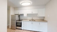This single-floor unit offers a well-organized layout comprising a combined kitchen and living area, a hallway, and a full bathroom — all finished with consistent light wood-look vinyl flooring that lends a clean, cohesive aesthetic throughout the space.

The kitchen is a functional standout, featuring crisp white shaker-style cabinetry extending both above and below the countertops, providing ample storage. Stainless steel appliances — including a gas range with oven and a full-size refrigerator — are neatly integrated alongside a double-basin sink. A flush-mount overhead light fixture provides even illumination, while a range hood above the stove adds practical ventilation.

The living area is open and bright, with a large multi-pane window fitted with horizontal blinds that invites natural light and frames a view of greenery outside. Baseboard heating units line the walls, ensuring comfortable climate control. Textured popcorn ceilings are consistent throughout, adding a characteristic finish to each room.

The hallway connects the main living areas to the bathroom, which is equipped with baseboard heating, a towel bar, and light neutral tones that carry through from the rest of the unit. The overall ambiance is clean, minimal, and move-in ready.