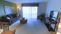This single-floor space centers around a well-appointed living room designed for comfort and practicality. The room features plush wall-to-wall carpeting that adds warmth underfoot, and is anchored by a large sliding glass door dressed with vertical blinds and a tailored fabric valance. The door allows an generous amount of natural light to fill the space while providing direct access to an outdoor area, visible through the glass. The living room is spaciously proportioned, comfortably accommodating a full-sized sofa adorned with decorative pillows, a woven round coffee table that introduces a natural texture, and a striped accent chair that adds visual interest. A tall floor lamp positioned near the sofa supplements the natural light during evening hours. The neutral color palette throughout — from the carpeting to the walls — creates a calm, versatile backdrop that suits a variety of furnishing styles. The overall ambiance is relaxed and inviting, with the interplay of natural light and soft furnishings contributing to a cozy yet open atmosphere well-suited for everyday living.