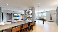 Located at 925 S Glebe Rd in Arlington, VA, this contemporary amenity lounge is a well-appointed, single-floor social space designed for both recreation and relaxation. The open-plan layout is anchored by a striking geometric room divider—a bold, black-framed shelving partition adorned with live greenery—that visually separates distinct activity zones while maintaining a cohesive, airy feel throughout.

The recreation area features a full-size billiards table set beneath a modern brass chandelier, complemented by a bold blue accent wall and curated wall art. Adjacent to it, a foosball table occupies a bright, windowed alcove with views of the surrounding Arlington neighborhood and a convenient wet bar nearby.

The lounge area offers a generously sized sectional sofa, organic-shaped coffee tables, and warm accent pillows arranged around a blue area rug, creating an inviting gathering space. A built-in fireplace with a wall-mounted television and integrated shelving adds a refined, residential warmth to this zone.

Light hardwood flooring runs throughout, unifying the space and enhancing the brightness provided by large windows on multiple walls. Recessed lighting and thoughtfully placed pendant and sconce fixtures ensure a well-lit, polished ambiance. Small café-style tables and chairs offer additional seating options, making this lounge a versatile and engaging communal destination.