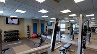 This fitness center offers a thoughtfully designed workout environment suited for a wide range of exercise routines. The space features a drop ceiling with recessed panel lighting that provides bright, even illumination throughout. Full-length mirrors line the walls, enhancing the sense of openness and allowing users to monitor their form during workouts. Natural light filters in through a window, adding to the welcoming atmosphere.

The facility is equipped with a multi-station cable and weight machine, a flat weight bench, and a dedicated free weights area with a tiered dumbbell rack offering a variety of weight options. Functional training equipment includes medicine balls displayed on a vertical rack, exercise stability balls, and ab training benches, catering to both strength and conditioning workouts. Rubberized flooring and exercise mats are laid throughout the space to provide safety and comfort underfoot.

A wall-mounted flat-screen television is positioned for easy viewing during cardio or stretching sessions. A blue accent door adds a subtle pop of color to the neutral beige walls. The overall layout is organized and spacious, ensuring a comfortable and efficient workout experience for users of varying fitness levels.