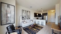 This stylish single-floor space showcases a thoughtfully designed open-concept layout that integrates the living and kitchen areas into one cohesive, functional environment. The living area is anchored by a light-toned sofa complemented by a sleek black accent chair, a round coffee table with geometric gold legs, and a bold navy and gold patterned area rug that adds warmth and visual interest to the light wood-style flooring. A striking arc floor lamp and a pair of large framed wall art pieces complete the sophisticated aesthetic. The kitchen is finished with dark espresso cabinetry that creates a dramatic contrast against the white peninsula countertop, which features a stone surface and comfortably seats multiple bar stools. Pendant lighting in brushed metal tones hangs above the peninsula, adding both task lighting and decorative flair. Stainless steel appliances, including a full-size refrigerator, contribute a polished, professional quality to the space. The neutral wall tones and light flooring throughout the open layout create a bright, airy ambiance, while the cohesive dark-and-light contrast in the design choices lends the space a modern, upscale character well-suited for both everyday living and entertaining.