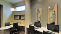 This single-floor business center offers a thoughtfully designed workspace suited for focused, professional use. The room features multiple individual workstations arranged along clean, white built-in desks, each equipped with desktop computers and keyboards, accommodating several users simultaneously. Ergonomic black office chairs provide comfortable seating throughout.

The space is finished with neutral gray-toned walls that create a calm, professional atmosphere. A standout decorative element is a tall mirror framed in natural live-edge wood, adding an organic warmth that contrasts pleasingly with the otherwise contemporary interior. Framed cityscape artwork adorns the walls, contributing a sophisticated, urban aesthetic.

Natural light filters in through a horizontal clerestory window positioned near the ceiling, brightening the space without causing screen glare. A modern pendant lamp provides additional ambient lighting. Dark tile flooring grounds the room and complements the overall neutral color palette.

The layout is efficiently organized, with a structural column subtly dividing the workspace into distinct areas, offering a degree of privacy between workstations while maintaining an open, cohesive feel throughout the room.