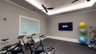 This dedicated fitness studio is thoughtfully designed to support an active lifestyle in a clean, comfortable environment. The room features neutral gray walls, crisp white crown molding, and dark carpet flooring — a practical combination that keeps the space looking sharp while standing up to regular use. A standout architectural detail is the tray ceiling, outfitted with integrated LED ambient lighting that creates a soft, even glow throughout the room, complemented by a sleek ceiling fan for airflow during intense workouts. Multiple stationary cycling bikes, including Pro Maxima and Silk models, are arranged to face a large wall-mounted flat-screen television that displays Fitness On Demand, offering users access to guided, on-demand workout classes at any time. A wide window with horizontal blinds allows natural light to filter into the space while maintaining privacy. Recessed ceiling speakers suggest built-in audio capability, enhancing the workout atmosphere. The overall ambiance is modern, functional, and focused — offering everything needed for an effective cardio session in a well-maintained, purpose-built environment.