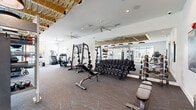This modern fitness center offers a comprehensive, single-floor layout thoughtfully designed to accommodate a wide range of workout disciplines. The space is divided into distinct functional zones, each outfitted with professional-grade equipment. The strength training area features a power rack with Olympic weights, multiple adjustable benches, and an extensive dumbbell rack, all positioned in front of large floor-to-ceiling mirrors that visually expand the room. A dedicated functional training zone includes a ceiling-mounted pull-up rig, a heavy punching bag, battle ropes, medicine balls, and TRX-style suspension trainers, enabling dynamic, high-intensity workouts. The cardio area lines the windowed perimeter, housing treadmills, ellipticals, stationary bikes, and a stair climber, all with views of outdoor palm trees and a pool area. Natural light floods through large windows along multiple walls, enhancing the open, airy atmosphere. Overhead, a striking decorative lattice ceiling installation adds an artistic warmth to the space, complemented by stylish pendant lighting and multiple ceiling fans for air circulation. The patterned carpet with teal accents runs throughout, tying the design together with a contemporary aesthetic. A mounted flat-screen television and wall art complete the polished, resort-style environment.