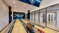 Located at 632 Newark Avenue in Jersey City, NJ, this contemporary amenity space seamlessly blends entertainment and relaxation into one well-appointed floor. The space is defined by two primary zones that flow together under recessed lighting and clean white ceilings. The bowling alley section features polished hardwood lanes, modern ball returns, large overhead scoring displays, and dramatic X-shaped neon lighting at the far end, all framed by sleek floor-to-ceiling glass partitions with dark steel frames. Adjacent to the lanes, an exposed brick accent wall adds industrial warmth and character, grounding the space with rich texture. The lounge area is generously furnished with large sectional sofas, a coffee table, and high-top bar seating positioned along expansive floor-to-ceiling windows that frame striking views of the Jersey City and Manhattan skylines. Natural light pours in throughout the day, brightening the light hardwood floors and cream-toned ceilings. Additional amenities visible within the space include a billiards table and wall-mounted shelving. The overall design balances modern industrial elements—exposed brick, steel, and glass—with comfortable, curated furnishings, creating a dynamic and inviting communal environment.