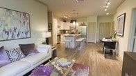 This thoughtfully designed single-floor residence offers an open-concept layout that seamlessly connects the living, dining, and kitchen areas into one cohesive and inviting space. The living area features a clean, contemporary aesthetic with light-toned walls, warm hardwood-style flooring, and modern furnishings, including a crisp white sofa accented with decorative pillows and a glass coffee table styled with tasteful décor.

The kitchen is a standout feature, boasting white cabinetry, dark granite countertops, and a full suite of stainless steel appliances, including a refrigerator, range, and built-in microwave. A central kitchen island with bar seating provides both additional prep space and a casual dining option, illuminated by elegant pendant lighting overhead. Track lighting along the kitchen ceiling adds a contemporary touch while ensuring the workspace is well-lit.

The dining area sits adjacent to the main living space and benefits from the open flow of the floor plan. Throughout the residence, consistent hardwood-style flooring ties each area together, enhancing the sense of continuity and spaciousness. The neutral color palette, modern light fixtures, and carefully chosen accents contribute to an overall ambiance that is both stylish and functional — ideal for everyday living and entertaining alike.