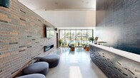 Located at 902 SE Brick Ave in Bentonville, Arkansas, this thoughtfully designed two-floor community space blends contemporary architecture with warm, inviting interiors across a generous footprint. The ground floor welcomes visitors with a dramatic lobby featuring soaring ceilings, a bold dark herringbone-tiled accent wall, polished concrete floors, and a sculptural reception desk. Adjacent to the lobby, a bright living lounge opens to sweeping floor-to-ceiling glass walls overlooking an outdoor pool area, anchored by an exposed brick fireplace wall with a wall-mounted TV. A coffee bar area features deep navy cabinetry, subway tile backsplash, and a communal high-top table on warm hardwood floors. A reading nook with large windows and a built-in magazine rack adds a relaxed, sun-filled retreat. The upper floor expands the space considerably, housing a striking lounge crowned by a vibrant large-scale "Love Your Neighbor" mural rising to double-height ceilings, complemented by an upstairs lounge with curated artwork. A library-style conference room features floor-to-ceiling bookshelves, a modern chandelier, a large wooden meeting table, and abundant natural light. A long communal work bar with task lighting and upholstered seating rounds out the upper level, making this a dynamic, community-centered destination.