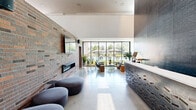 Located at 902 SE Brick Ave in Bentonville, Arkansas, this thoughtfully designed two-floor community space blends contemporary architecture with warm, inviting interiors across a generous footprint. The ground floor welcomes visitors with a dramatic lobby featuring soaring ceilings, a bold dark herringbone-tiled accent wall, polished concrete floors, and a sculptural reception desk. Adjacent to the lobby, a bright living lounge opens to sweeping floor-to-ceiling glass walls overlooking an outdoor pool area, anchored by an exposed brick fireplace wall with a wall-mounted TV. A coffee bar area features deep navy cabinetry, subway tile backsplash, and a communal high-top table on warm hardwood floors. A reading nook with large windows and a built-in magazine rack adds a relaxed, sun-filled retreat. The upper floor expands the space considerably, housing a striking lounge crowned by a vibrant large-scale "Love Your Neighbor" mural rising to double-height ceilings, complemented by an upstairs lounge with curated artwork. A library-style conference room features floor-to-ceiling bookshelves, a modern chandelier, a large wooden meeting table, and abundant natural light. A long communal work bar with task lighting and upholstered seating rounds out the upper level, making this a dynamic, community-centered destination.