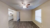 This 2-floor space is designed with a functional and straightforward layout. The main living area features dark hardwood-style flooring that contrasts with the warm beige walls, creating a clean and inviting atmosphere. A ceiling fan with an integrated light fixture is centrally mounted on the textured popcorn ceiling, providing both airflow and ambient lighting. A window fitted with blinds allows natural light to filter into the room, brightening the space throughout the day. A floor vent is also visible, indicating a built-in HVAC system for climate control.

An open doorway connects the living area to adjacent spaces, offering a glimpse into a kitchen outfitted with white cabinetry and what appears to be a countertop with a sink. The white baseboards throughout the space add a crisp finishing detail against the dark flooring. The overall layout feels open and versatile, suitable for various furniture arrangements. The combination of practical finishes, natural light, and functional features makes this a well-appointed space across its two floors.