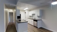 This single-floor space presents a clean, modern aesthetic highlighted by a thoughtfully designed kitchen at its heart. The kitchen features crisp white flat-panel cabinetry that extends to the upper walls, providing ample storage, complemented by bold dark countertops that create a striking contrast. A peninsula with a matching dark countertop extends into the living area, offering additional prep space and an informal gathering point. Stainless steel appliances, including a dishwasher and refrigerator, add a contemporary touch, while a white subway tile backsplash brings subtle texture to the space. A stainless steel sink with a modern faucet is neatly integrated beneath a flush-mounted ceiling light that ensures the workspace is well-lit. Wood-look plank flooring runs throughout, adding warmth and visual continuity across the open layout. The neutral grey and white color palette creates a bright, airy ambiance, while the open sightlines connecting the kitchen to the hallway and adjoining rooms contribute to an overall sense of spaciousness. The layout is practical and well-organized, making efficient use of every area within this single-level residence.