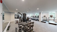 Located at 1435 51st Street in North Bergen, NJ, this fitness center offers a thoughtfully designed, single-floor space that provides a comprehensive workout environment. The gym features a clean, modern aesthetic with light gray walls, recessed lighting, and durable carpet flooring throughout.

The cardio section is lined with a row of treadmills, elliptical machines, and stationary cycling bikes from the True brand, positioned along large windows that frame views of lush greenery — creating a bright and invigorating atmosphere for exercise.

The strength training area is well-appointed with a multi-station cable machine, adjustable benches, and a full rack of dumbbells in graduated weights, making it suitable for a wide range of resistance workouts. A floor-to-ceiling mirror spans one wall, enhancing the sense of openness and allowing users to monitor their form during training.

A dedicated functional fitness zone features a power tower for bodyweight exercises, medicine balls, stability balls, and resistance accessories neatly organized on wall-mounted racks. Black-framed glass doors provide entry access and contribute to the facility's polished, contemporary look. The space benefits from consistent natural light through multiple windows, offering views of surrounding trees and creating a pleasant, energizing environment for all fitness levels.
