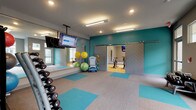 This well-appointed fitness center is organized across a single floor into distinct functional zones, offering a comprehensive workout environment. The space is unified by a bold teal and gray color palette applied to the walls, creating an energetic yet cohesive atmosphere throughout.

The main exercise floor features neutral carpet tiles interspersed with teal accent panels, providing cushioned, versatile workout surfaces. A full dumbbell rack, cable machines, a power rack with bench, and medicine balls with stability balls are among the equipment available, catering to a wide range of strength and functional training routines. Wall-mounted and ceiling-mounted flat-screen televisions are positioned throughout, ensuring visibility from multiple workout stations.

A separate spin studio is accessible through distinctive herringbone-patterned barn-style sliding doors. This room features hardwood flooring, a row of stationary cycling bikes, a ceiling-mounted television, and a large wall mirror that reflects natural light from tall windows along one side, creating a bright and spacious feel.

Throughout the facility, recessed and linear LED lighting provides consistent illumination, while large windows in key areas bring in natural light and exterior views. The overall design balances functionality with a modern, polished aesthetic suited for active daily use.