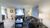 This single-floor space showcases a thoughtfully designed living room that balances comfort and style. The room is anchored by a pair of deep charcoal-blue sofas arranged around a sleek black coffee table with a mirrored surface, creating an inviting central seating area. A striped area rug in coordinating tones defines the space and adds visual texture underfoot.

Natural light filters into the room through a central window dressed with layered curtains — sheer white panels paired with elegant blue drapes — lending the space a soft, airy quality. Neutral-toned walls provide a calm backdrop that allows the furnishings and décor to take center stage.

A large landscape painting in cool blue and grey tones commands attention on one wall, complementing the overall color palette. Decorative accents, including an oversized wall clock, sculptural pieces, and throw pillows in earthy neutrals, add personality and depth to the room. Warm lamplight contributes to a cozy atmosphere, making this living space both functional and visually cohesive throughout.