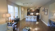 This thoughtfully designed single-floor space showcases an open-concept layout that seamlessly connects the living and kitchen areas, creating a spacious and cohesive environment. Rich hardwood flooring flows throughout, anchoring the warm, neutral color palette of the walls and trim.

The kitchen is a standout feature, appointed with dark espresso cabinetry that extends to the ceiling, under-cabinet lighting, a tile backsplash, and black appliances including a refrigerator and range. A central white kitchen island doubles as a breakfast bar, accommodating three adjustable bar stools — making it ideal for casual dining or entertaining.

Pendant lighting hangs above the island, while a track lighting system illuminates the living area, offering both ambient and directional light. Large windows along the exterior wall flood the space with natural light and offer views of the surrounding exterior, further enhancing the open, airy atmosphere.

The living area is generously proportioned, with ample room for comfortable furnishings. A decorative console table and curated accessories add character to the space. The overall ambiance is modern yet inviting, striking a balance between functional design and stylish aesthetics suited for everyday living.