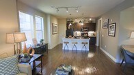 This thoughtfully designed single-floor space showcases an open-concept layout that seamlessly connects the living and kitchen areas, creating a spacious and cohesive environment. Rich hardwood flooring flows throughout, anchoring the warm, neutral color palette of the walls and trim.

The kitchen is a standout feature, appointed with dark espresso cabinetry that extends to the ceiling, under-cabinet lighting, a tile backsplash, and black appliances including a refrigerator and range. A central white kitchen island doubles as a breakfast bar, accommodating three adjustable bar stools — making it ideal for casual dining or entertaining.

Pendant lighting hangs above the island, while a track lighting system illuminates the living area, offering both ambient and directional light. Large windows along the exterior wall flood the space with natural light and offer views of the surrounding exterior, further enhancing the open, airy atmosphere.

The living area is generously proportioned, with ample room for comfortable furnishings. A decorative console table and curated accessories add character to the space. The overall ambiance is modern yet inviting, striking a balance between functional design and stylish aesthetics suited for everyday living.