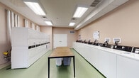 Located at 115 Carriage Hill Dr in Athens, Ohio, this single-floor shared laundry facility is designed to be both functional and comfortable for everyday use. The main laundry area is lined with a full row of top-load washing machines along one wall and a bank of stacked dryers along the opposite side, all finished in white and equipped with modern payment terminals. Overhead, recessed fluorescent panel lighting illuminates the space evenly, while a light green floor and warm peach-toned walls create a clean, welcoming environment. A large butcher-block folding table anchors the center of the room, providing ample space for sorting and folding laundry. A row of white storage lockers offers secure personal storage for residents. Adjacent to the laundry area, a dedicated lounge corner adds a comfortable waiting space, furnished with leather and upholstered armchairs, a zebra-print area rug, a wall-mounted flat-screen television, and sheer white curtains framing the windows. Deep green accent walls give the lounge a stylish, modern feel. Snack and beverage vending machines are conveniently positioned nearby, rounding out the amenities and making this facility a practical, all-in-one community resource.