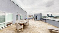 This expansive rooftop terrace offers a well-appointed outdoor living experience elevated above a dense urban landscape. The floor is covered throughout in light-toned interlocking wood deck tiles, lending a clean, contemporary aesthetic to the open-air space. A low perimeter wall topped with glass panels runs along the outer edges, preserving unobstructed panoramic views of the surrounding city skyline while maintaining a sense of security.

The terrace is organized into distinct functional zones. A generous lounge area features a large wraparound wicker sectional sofa paired with a matching coffee table, positioned to face the cityscape. Nearby, a set of reclining chaise lounges provides additional relaxation options. A separate dining zone accommodates a rustic wood table surrounded by wicker chairs, suitable for outdoor meals or gatherings.

A central rooftop access structure with a glazed door serves as the primary entry point, flanked by exterior lighting and a fire alarm. Additional access is available through a door set into the building's gray stucco facade, which also features a frosted-glass window. Waste receptacles are discreetly placed near the perimeter. The overall layout is open and versatile, with ample circulation space throughout.