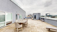 This expansive rooftop terrace offers a well-appointed outdoor living experience elevated above a dense urban landscape. The floor is covered throughout in light-toned interlocking wood deck tiles, lending a clean, contemporary aesthetic to the open-air space. A low perimeter wall topped with glass panels runs along the outer edges, preserving unobstructed panoramic views of the surrounding city skyline while maintaining a sense of security.

The terrace is organized into distinct functional zones. A generous lounge area features a large wraparound wicker sectional sofa paired with a matching coffee table, positioned to face the cityscape. Nearby, a set of reclining chaise lounges provides additional relaxation options. A separate dining zone accommodates a rustic wood table surrounded by wicker chairs, suitable for outdoor meals or gatherings.

A central rooftop access structure with a glazed door serves as the primary entry point, flanked by exterior lighting and a fire alarm. Additional access is available through a door set into the building's gray stucco facade, which also features a frosted-glass window. Waste receptacles are discreetly placed near the perimeter. The overall layout is open and versatile, with ample circulation space throughout.