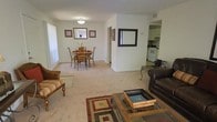 This single-floor residence presents an open-concept layout that seamlessly connects the living, dining, and kitchen areas into one cohesive living space. The living area is anchored by a glass-top coffee table and features a mix of seating, including a leather armchair and a striped accent chair, all resting on a patterned area rug over wall-to-wall carpeting. Neutral walls and light-toned carpet throughout contribute to a bright, airy ambiance.

The dining area is positioned adjacent to the living space, accommodating a round wooden dining table with cushioned chairs and decorated with framed artwork and a small floral centerpiece. A flush-mount ceiling light fixture provides direct illumination to the dining space.

A partial wall with a framed pass-through window separates the main living area from the kitchen, which is visible in the background. The kitchen features white cabinetry, a white range, and appears compact yet functional with visible counter space and appliances.

Natural light filters in through windows on the left side of the living room, enhancing the overall warmth of the space. The layout is practical and well-organized, making efficient use of the available floor space.