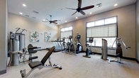 This single-floor fitness center is thoughtfully designed to support a full range of workout routines in a clean, modern environment. The space is outfitted with a comprehensive selection of cardio equipment, including multiple treadmills, elliptical machines, and stationary bikes, all arranged to allow comfortable movement throughout the room. Strength training is also well-represented, with an adjustable workout bench and additional free-weight equipment available for resistance training.

The room features a neutral color palette with light-toned walls and carpeted flooring, contributing to a calm and focused atmosphere. Two ceiling fans are mounted overhead, ensuring consistent airflow and comfort during intense workouts. Recessed lighting supplements the abundant natural light that streams in through a series of large, high-mounted windows fitted with adjustable shades, allowing users to control light levels as needed.

Decorative wall art adds a touch of personality to the space without detracting from its functional purpose. The overall layout is open and spacious, providing ample room between equipment stations for safe and comfortable use. This fitness center is well-suited for individuals seeking a clean, fully equipped environment for their daily exercise routine.